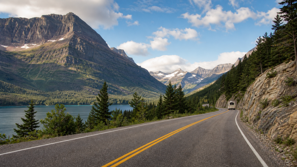 Scenic Glacier National Park road curving past an alpine lake toward rugged Montana mountain peaks with a small RV in the distance.
